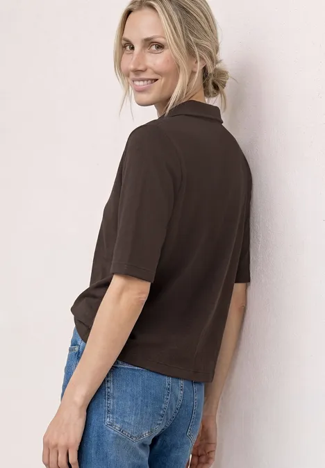 Person seitlich von hinten in dunkelbraunem Poloshirt und blauer Jeans, Haare zum Zopf gebunden.